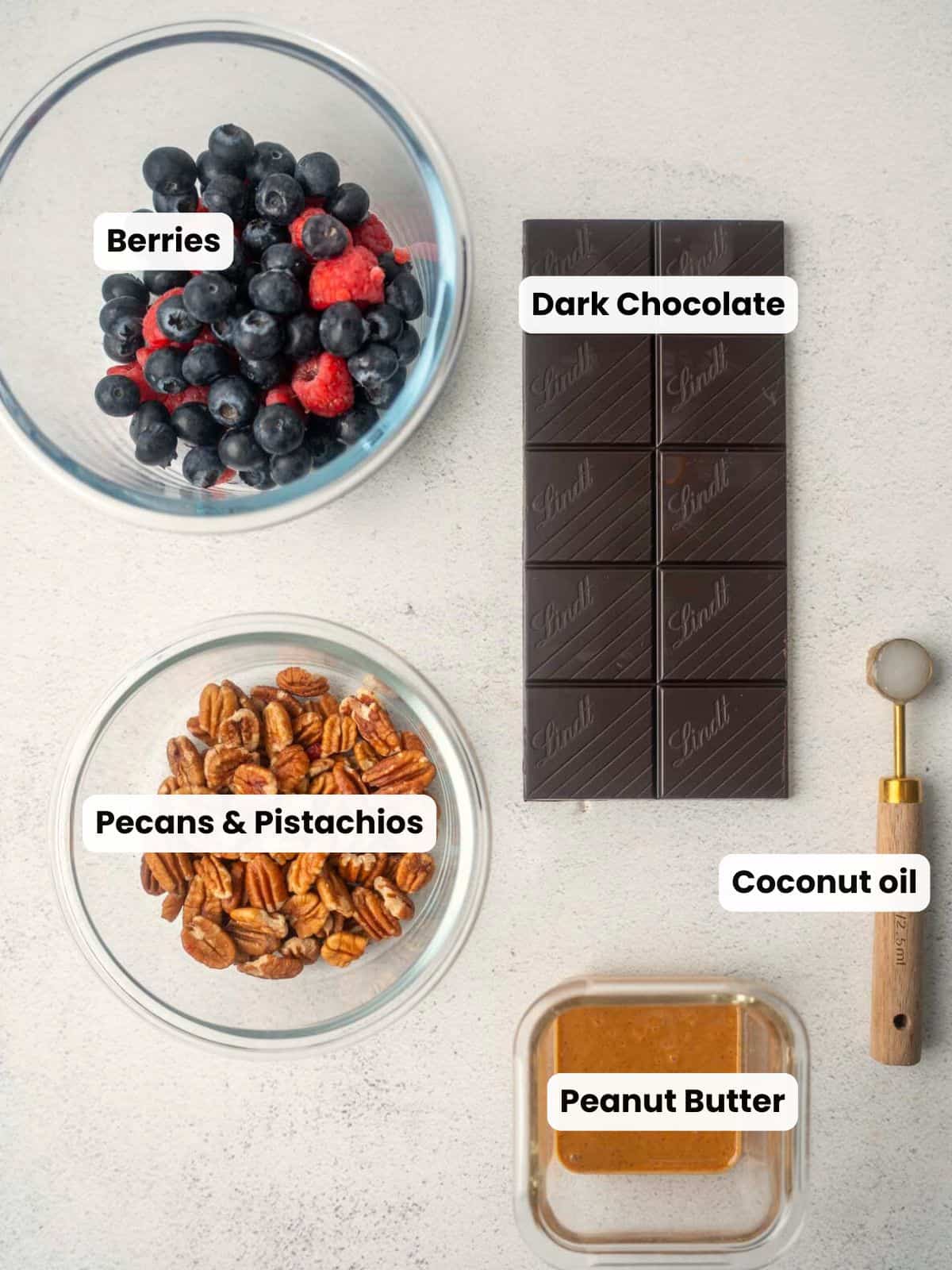 Overhead view of ingredients for healthy rocky road, including bowls of mixed berries, pecans and pistachios, a bar of dark chocolate, a small glass container of peanut butter, and a measuring spoon with coconut oil on a light surface.
