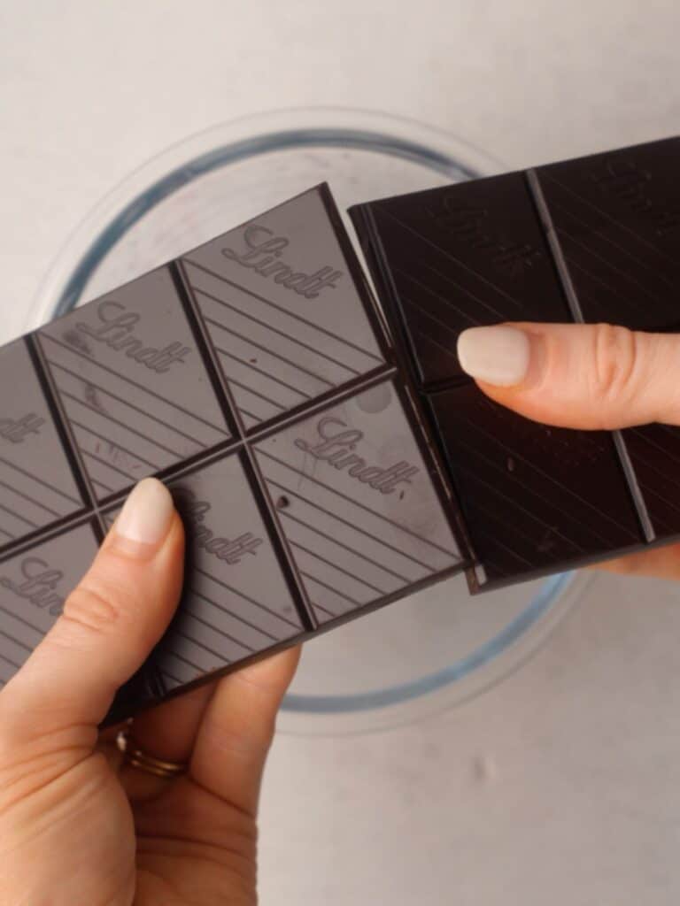 Hands breaking a bar of Lindt dark chocolate over a bowl in preparation for melting.