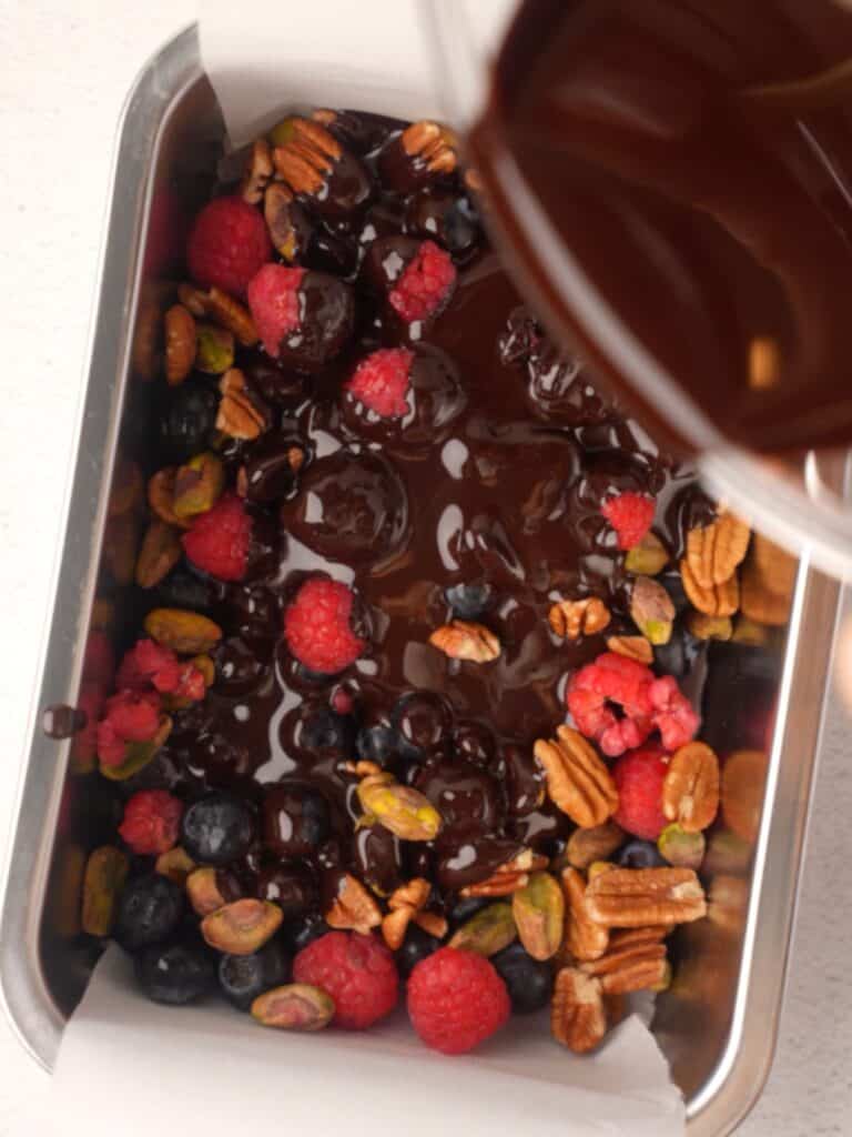 Pouring melted dark chocolate over a mixture of fresh raspberries, blueberries, pecans, and pistachios in a parchment-lined loaf pan.