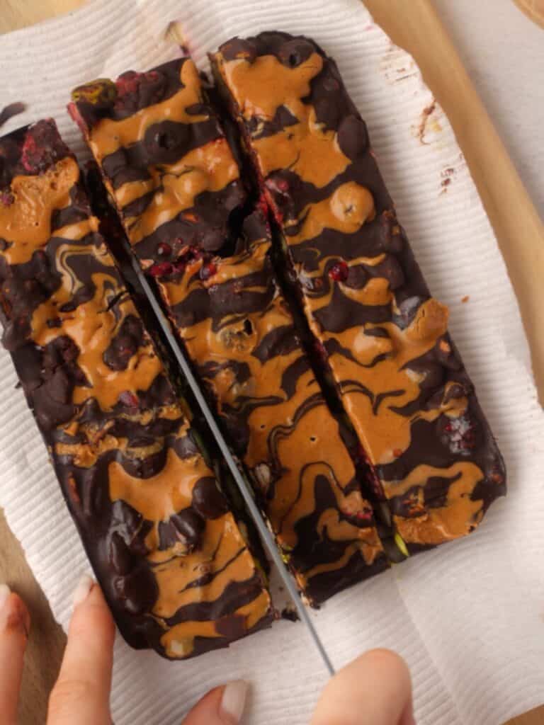 Overhead view of hands slicing a block of dark chocolate rocky road drizzled with peanut butter on a wooden board lined with parchment paper.