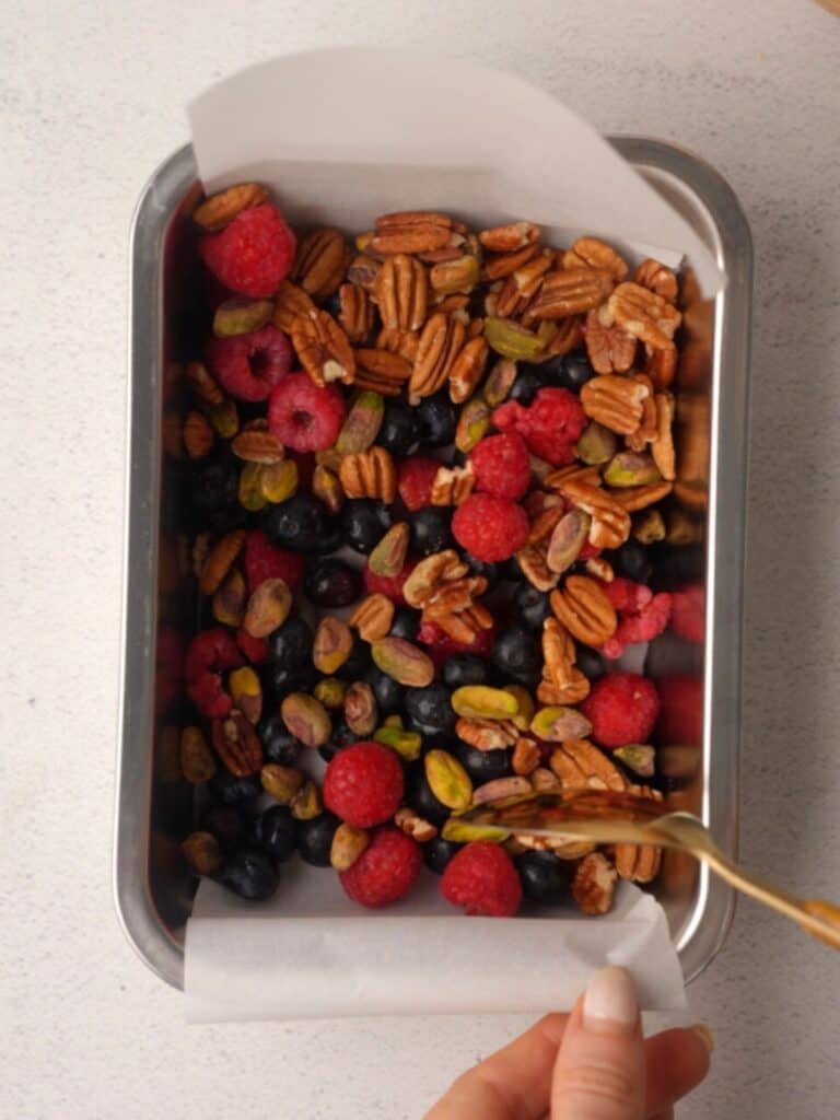 Parchment-lined loaf pan filled with raspberries, blueberries, pecans, and pistachios, ready for the next step in making healthy rocky road.