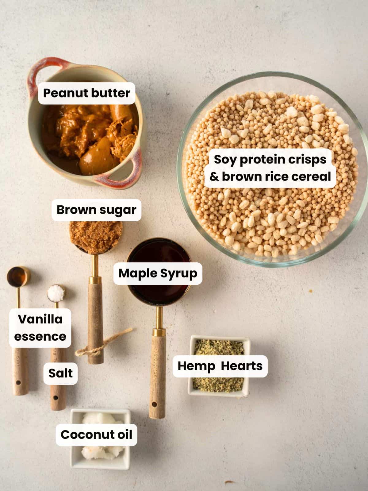 Overhead photo of ingredients for protein crisp bars including peanut butter, soy protein crisps, brown rice cereal, maple syrup, brown sugar, coconut oil, hemp hearts, vanilla, and salt arranged on a light countertop.