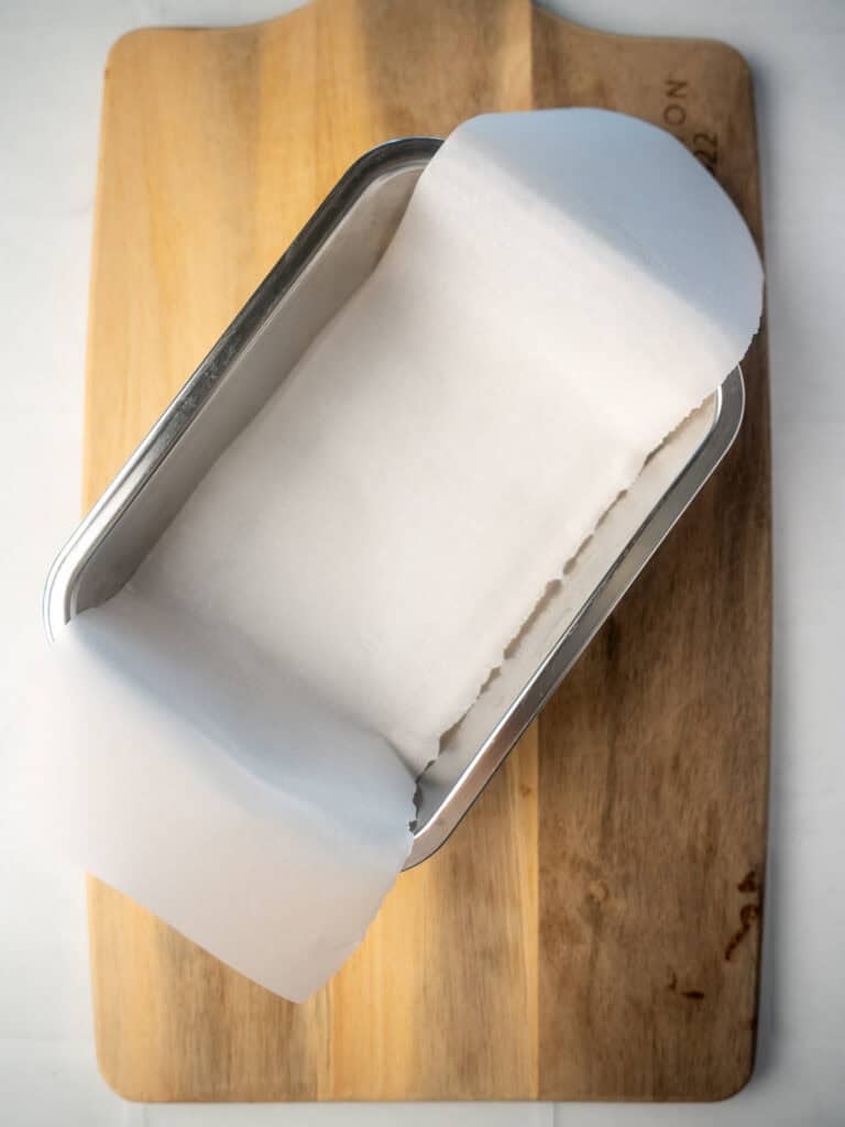 Loaf pan lined with parchment paper, placed on a wooden cutting board, ready for making healthy rocky road or another dessert.