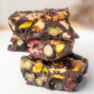 Stack of homemade dark chocolate healthy rocky road pieces filled with colorful nuts and fruit on a white surface.