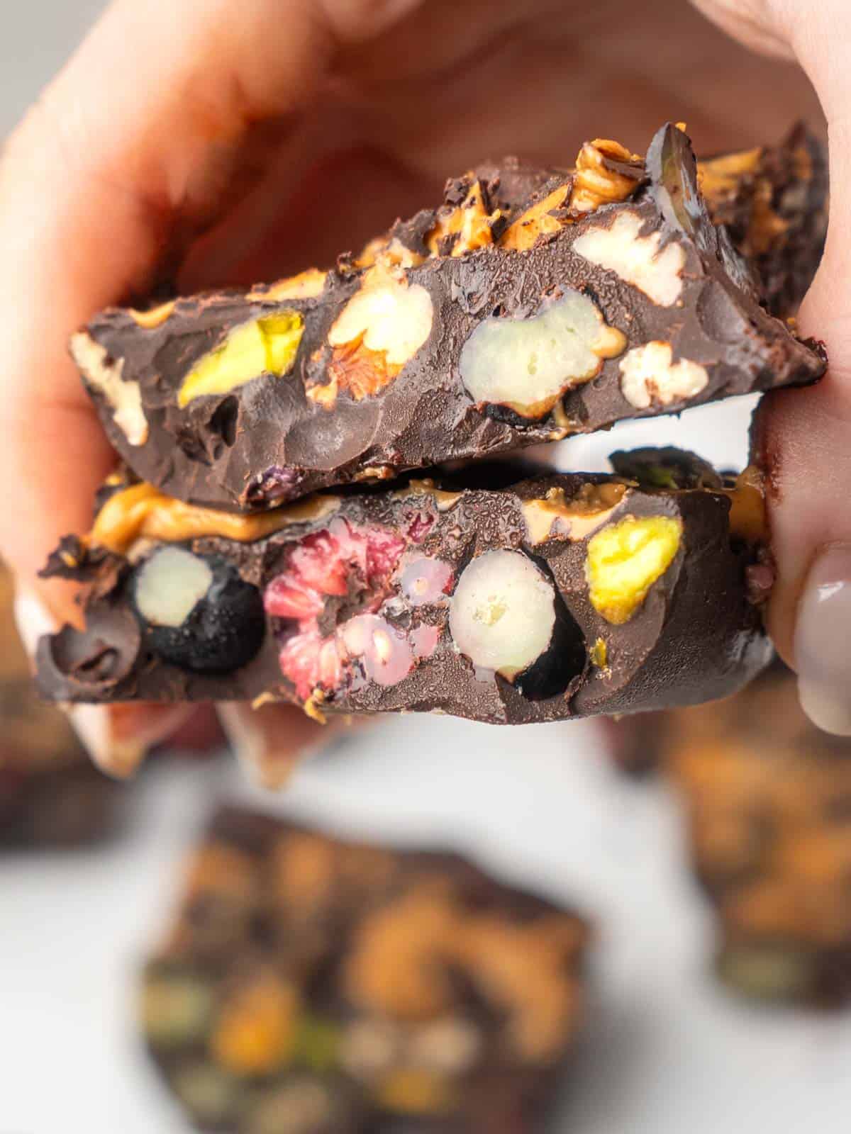 Close-up of a hand holding two pieces of dark chocolate healthy rocky road filled with nuts, pistachios and berries.