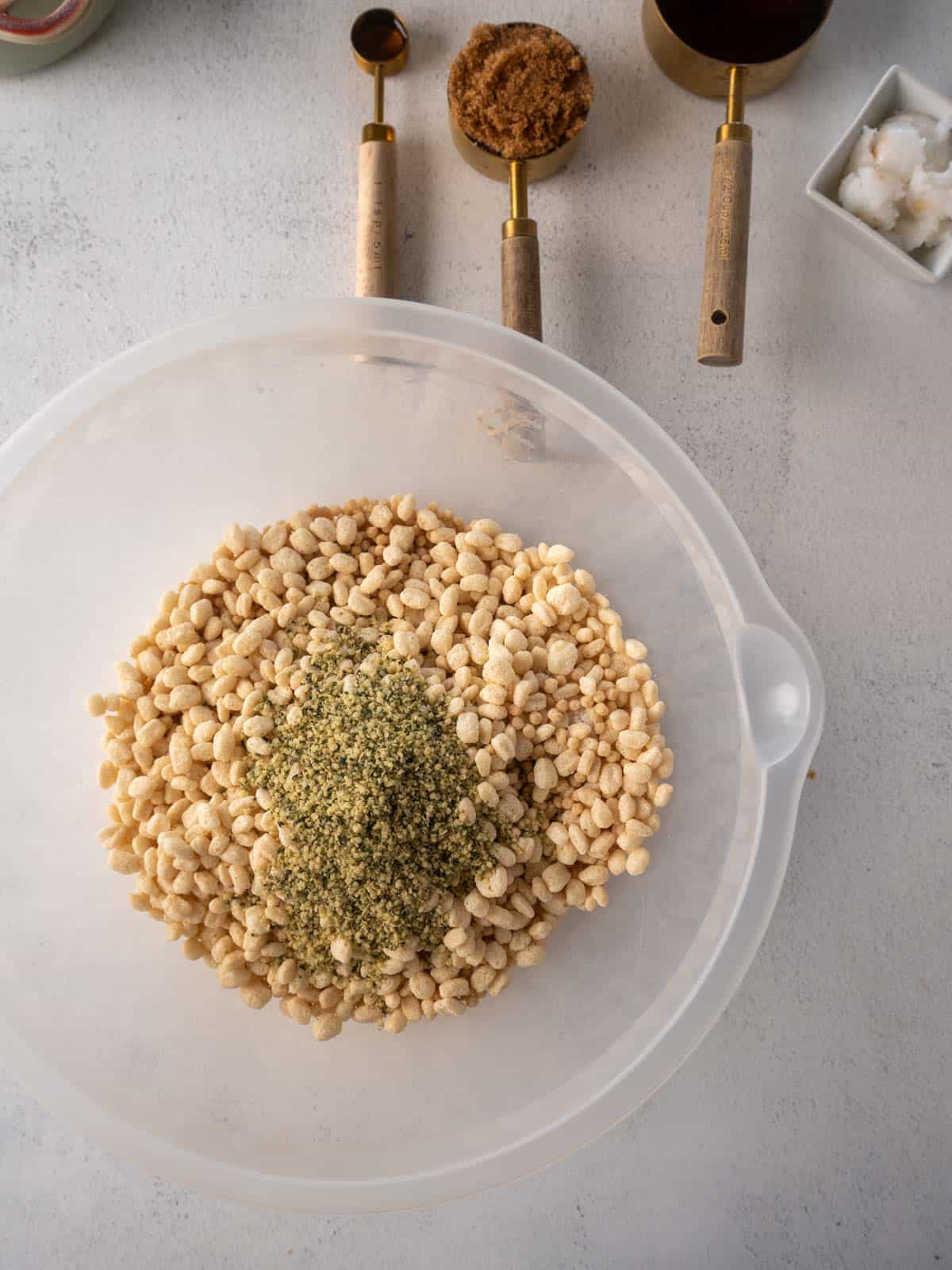 Overhead shot of soy protein crisps and hemp hearts in a mixing bowl, with measuring cups of brown sugar, maple syrup, and coconut oil in the background.