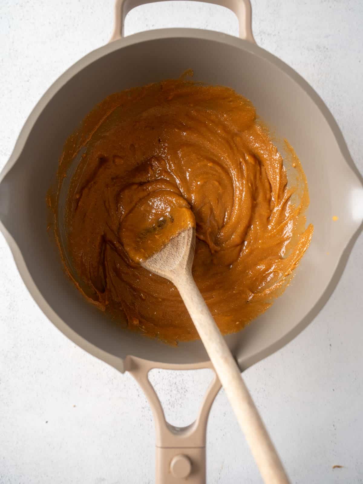 Peanut butter, maple syrup, coconut oil, and brown sugar melted together in a saucepan, being stirred with a wooden spoon until smooth.