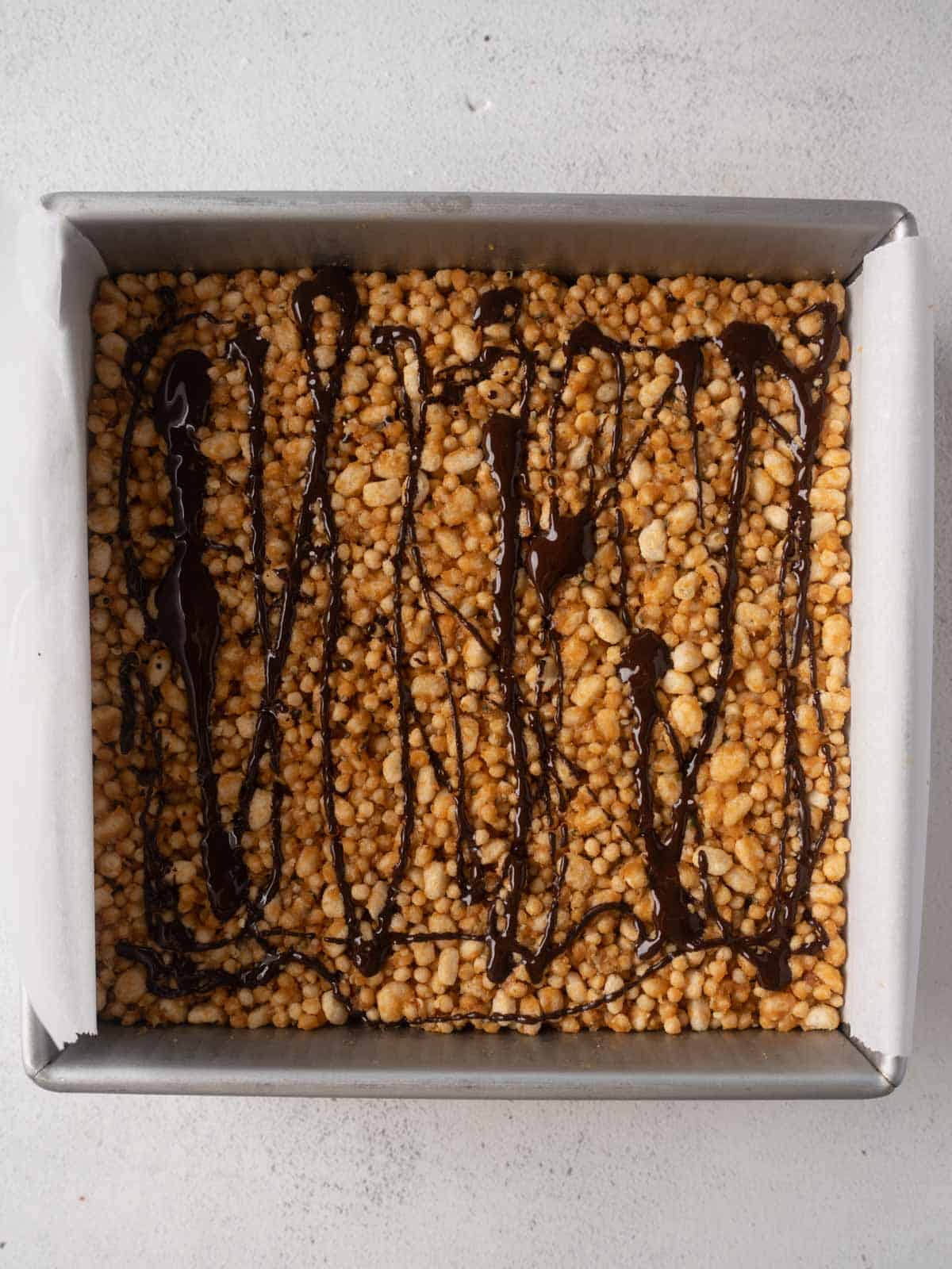 Protein crisp mixture pressed into a lined square baking pan and drizzled with melted dark chocolate.