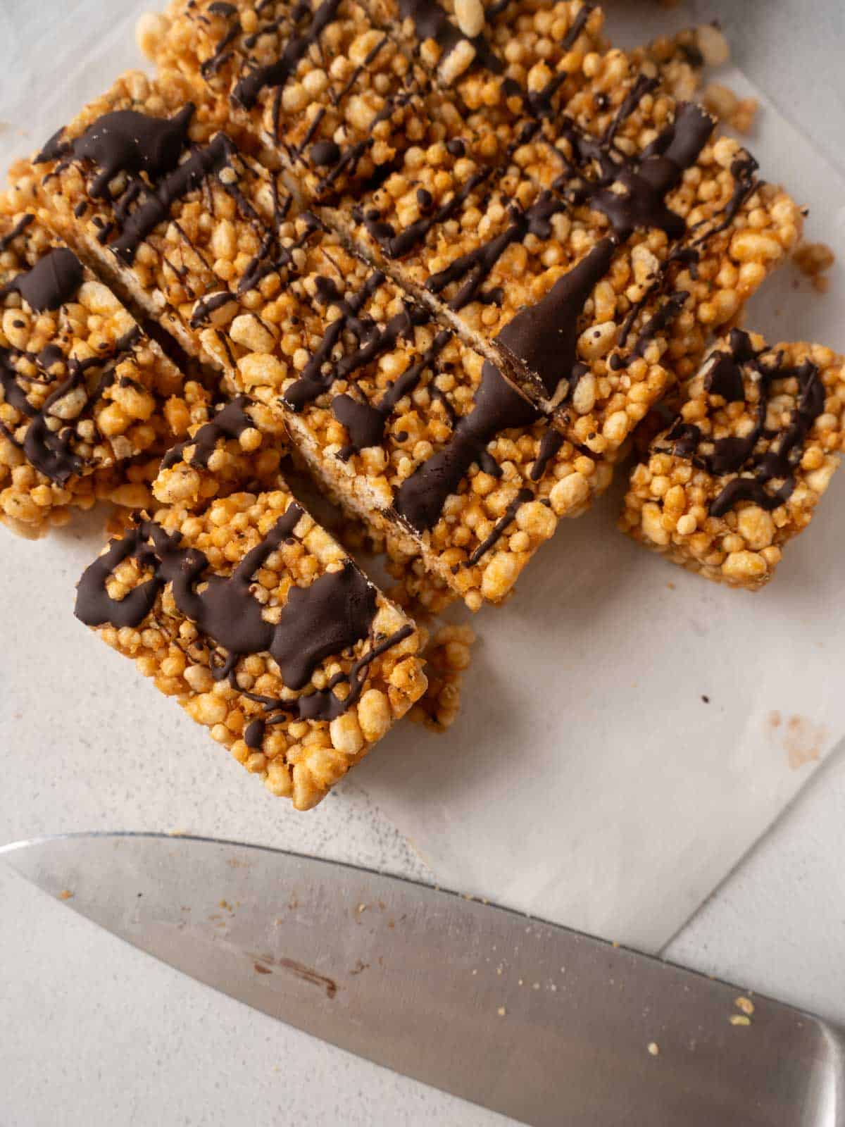 Protein crisp bars cut into squares on parchment paper, drizzled with dark chocolate, with a knife resting beside them.
