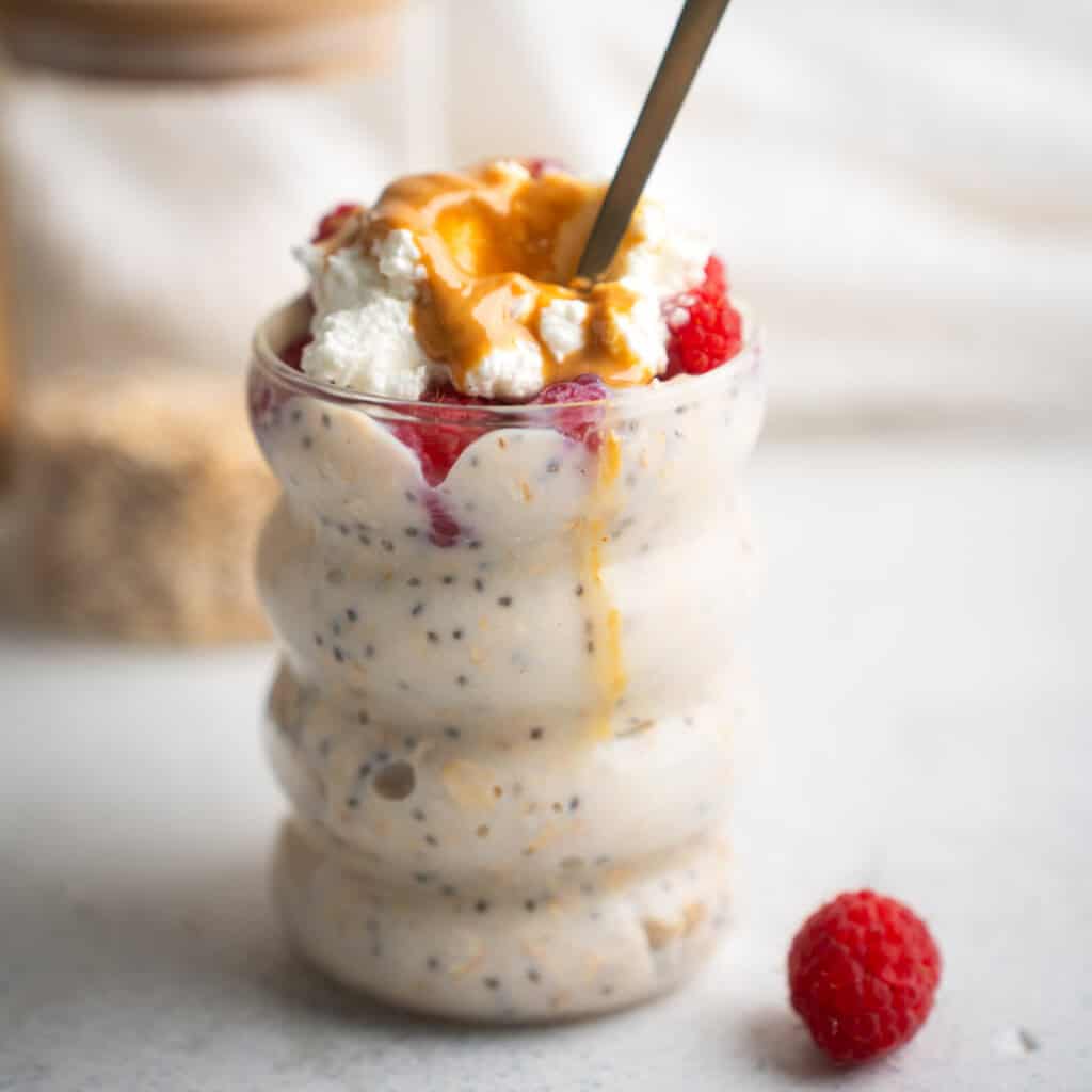 high protein overnight oats in a glass jar with a spoon sticking out and a raspberry in the foreground.