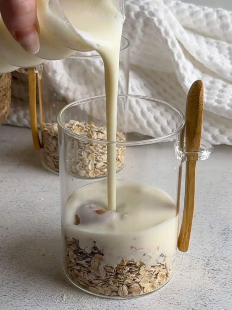 Milk being poured into a glass jar filled with oats, sitting on a light surface with a wooden-handled spoon resting on the side of the jar.