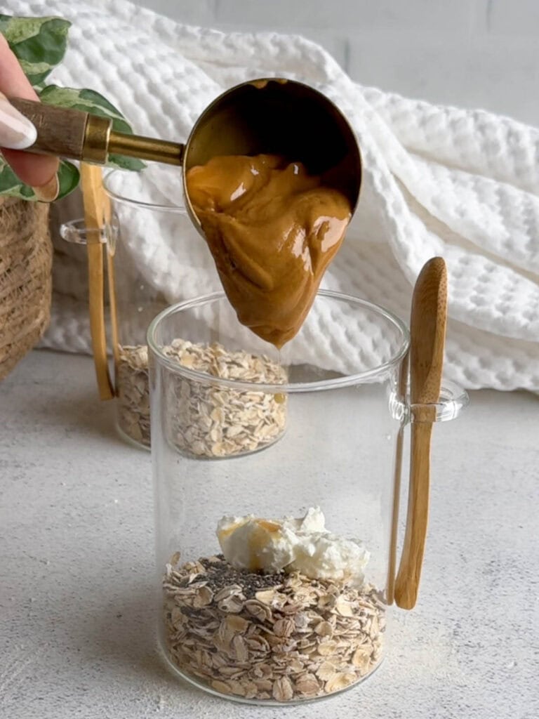 A scoop of creamy peanut butter being added to a glass jar containing oats, chia seeds, and Greek yogurt, with a wooden-handled spoon attached to the jar.