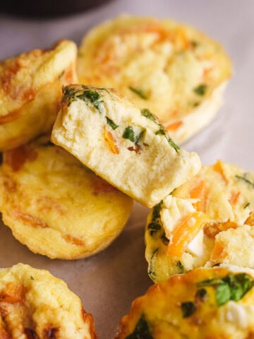 Golden brown egg bites filled with spinach, cheese, and vegetables, stacked on parchment paper with one cut open to show the fluffy texture inside.
