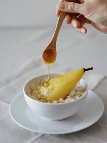 An anti-inflammatory recipe featuring a poached pear with honey served over creamy oatmeal in a white bowl, with a hand drizzling honey from a wooden spoon.