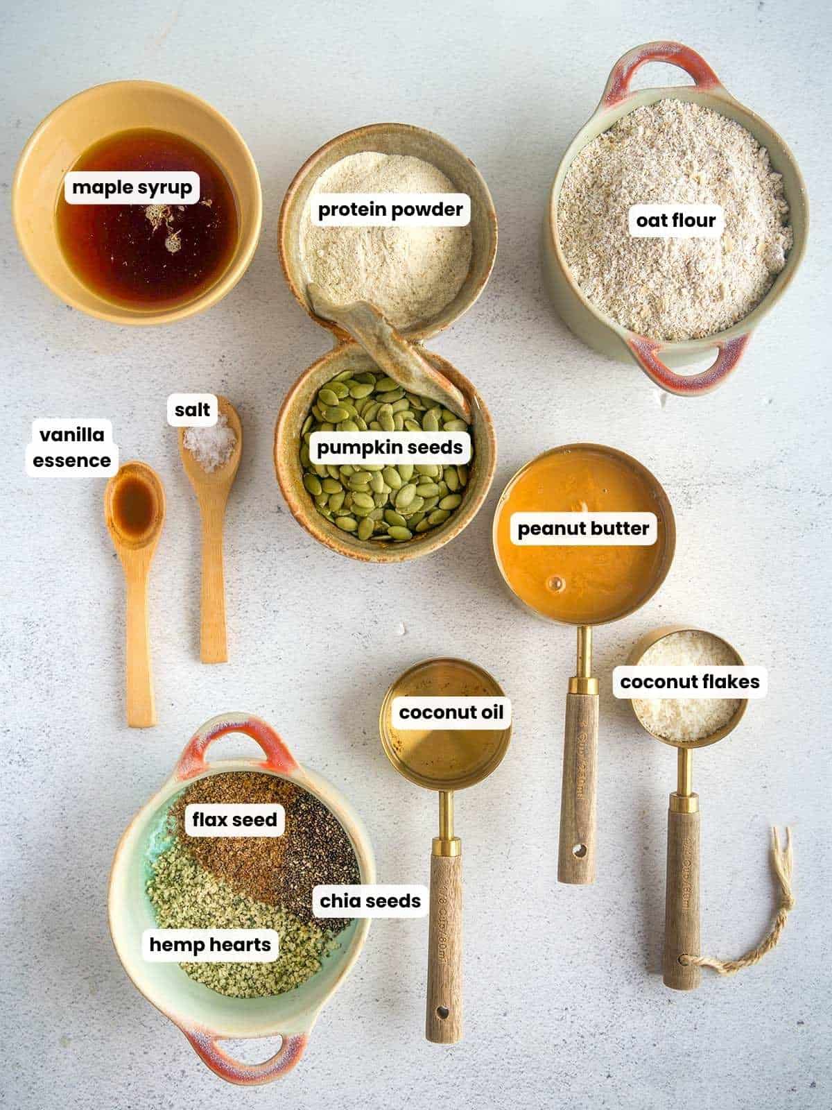 Overhead view of measured ingredients for oat breakfast bars, including bowls of oat flour, protein powder, peanut butter, maple syrup, pumpkin seeds, flaxseed, chia seeds, hemp hearts, coconut flakes, coconut oil, vanilla essence, and salt, arranged neatly on a light background.