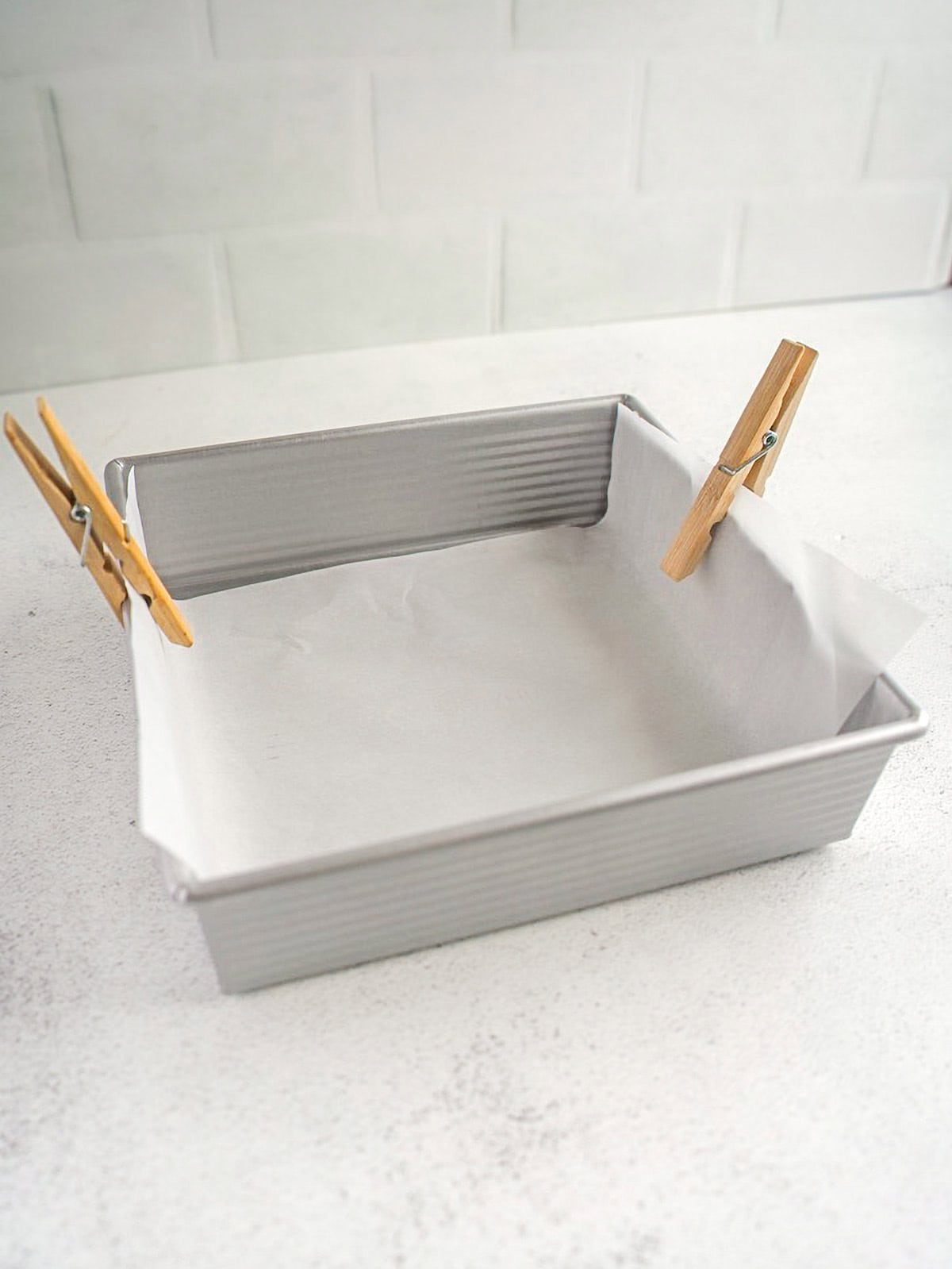 Square baking pan lined with parchment paper, with two wooden clothespins holding the paper in place.