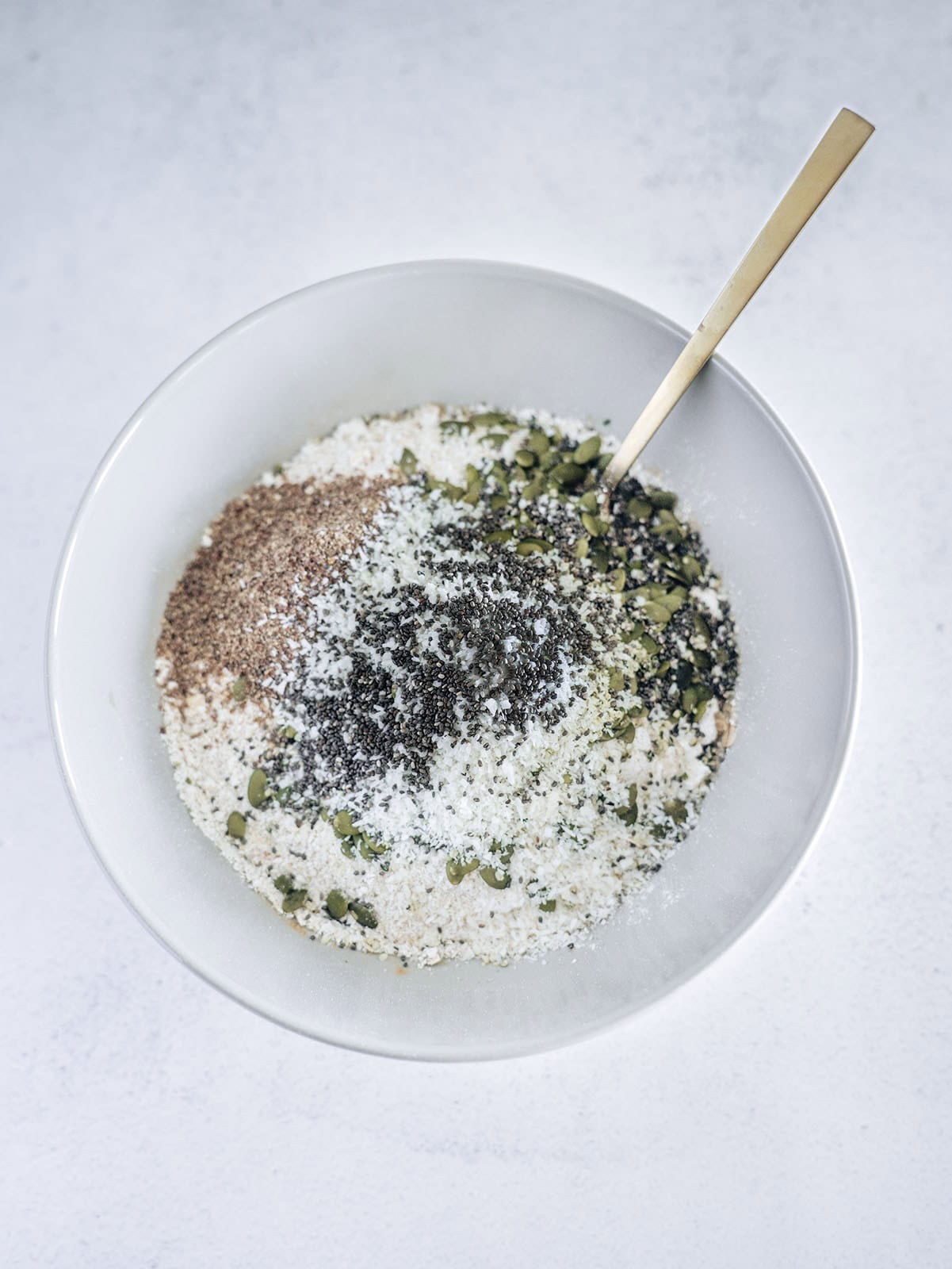 Mixing bowl filled with dry ingredients for oat breakfast bars, including oats, pumpkin seeds, chia seeds, flaxseed, and coconut flakes, before combining with wet ingredients.