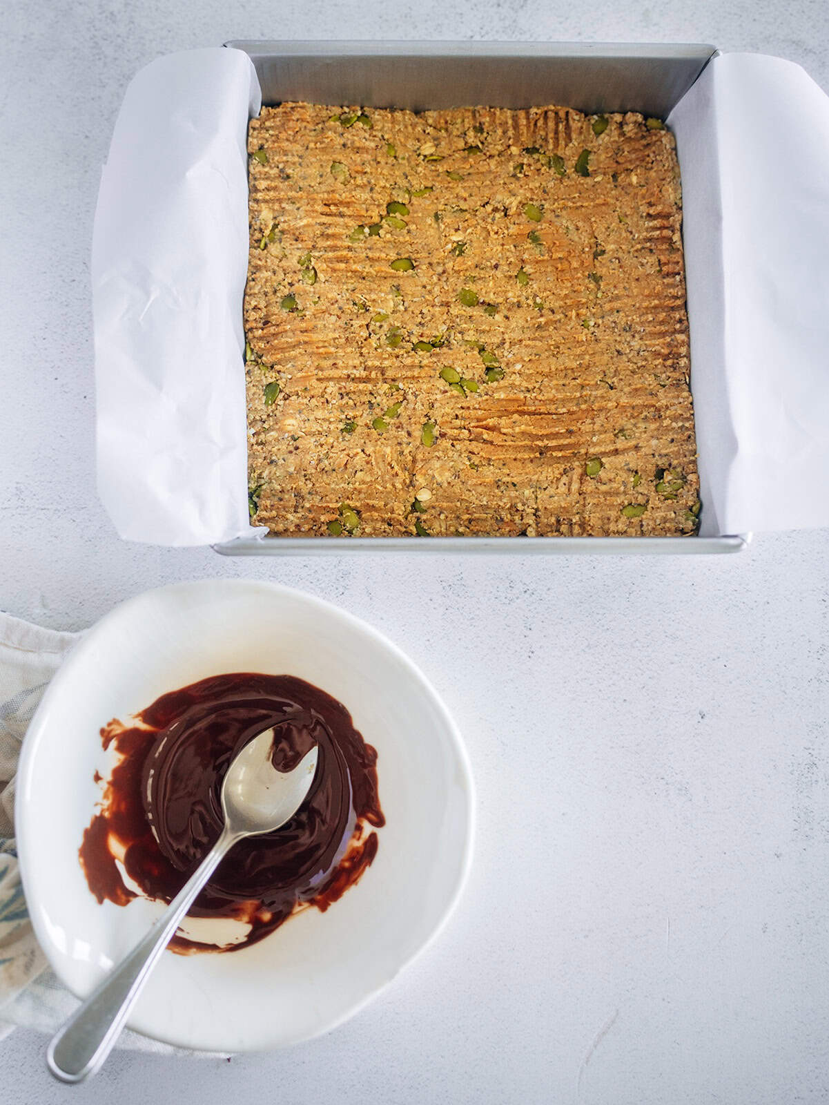 Baking pan filled with oat breakfast bar mixture beside a bowl of melted chocolate with a spoon, ready for drizzling.