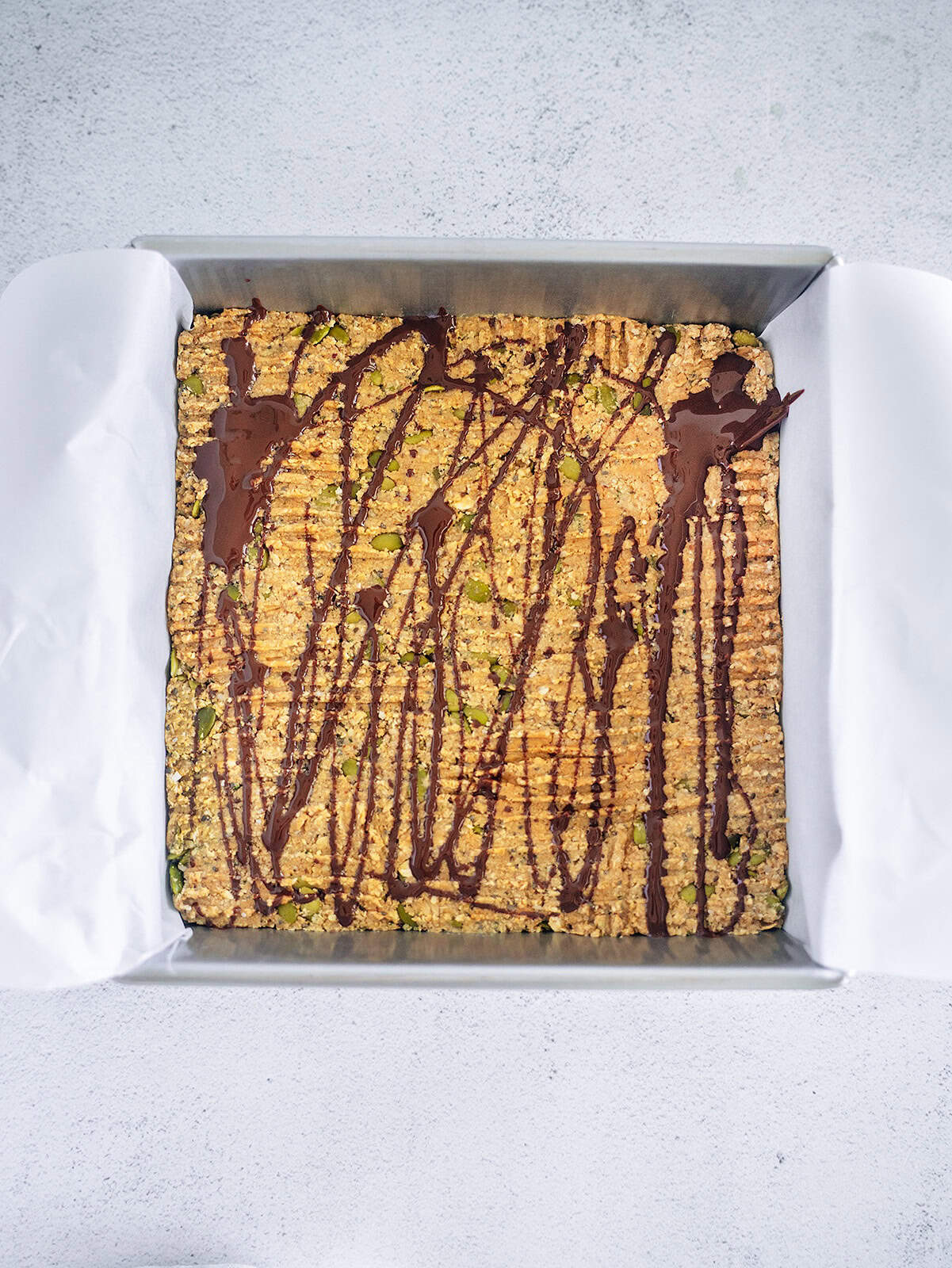Oat breakfast bar mixture pressed into a square baking pan, topped with melted chocolate drizzle, ready to set.