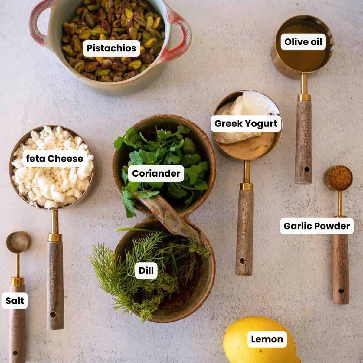 Overhead view of ingredients for a feta pistachio dip, including pistachios, feta cheese, Greek yogurt, olive oil, lemon, dill, coriander, garlic powder, and salt arranged neatly on a light surface with labeled measuring cups.