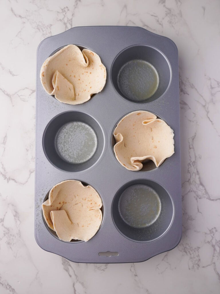 Mini tortillas pressed into four cups of a six-hole muffin tin, ready to be filled with the egg mixture.