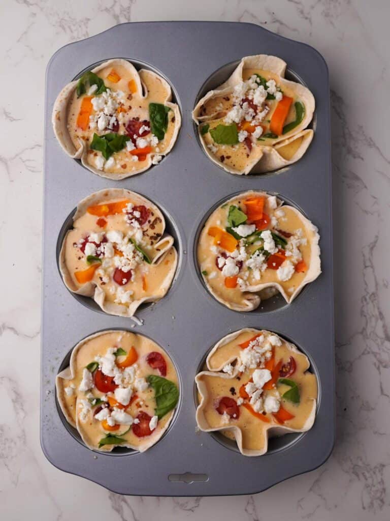 Tortilla egg cups filled with egg, cottage cheese, spinach, tomatoes and feta in a six-hole muffin tray, ready to bake.