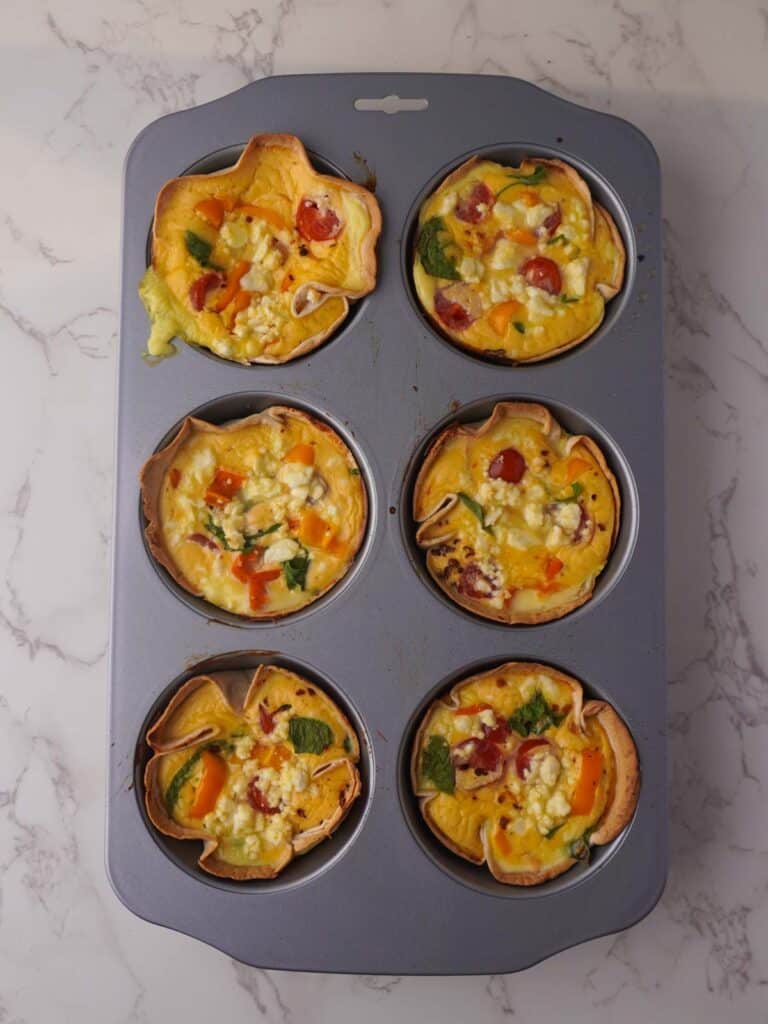 Baked tortilla egg cups with golden, crisp edges and a colorful egg, spinach, tomato and feta filling in a six-hole muffin tray.