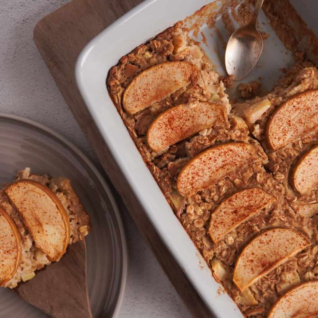 Baked oats made with apples served in a baking dish with one slice removed and laying on a plate nearby.