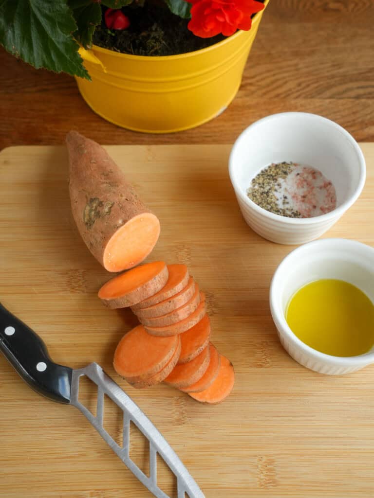 Fresh sweet potato being sliced on a wooden cutting board with olive oil and seasoning on the side.