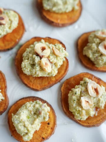 Baked sweet potato slices topped with a creamy green pesto-like spread and garnished with chopped hazelnuts, arranged on a white plate.
