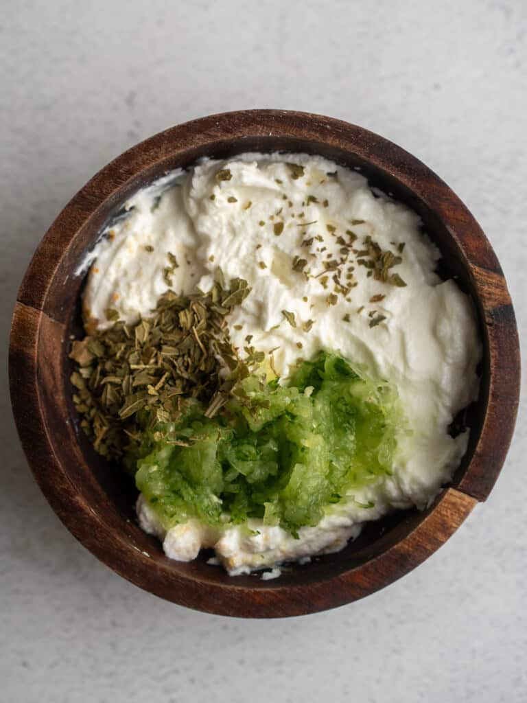 Wooden bowl with Greek yogurt, shredded cucumber, and dried herbs before being mixed into homemade tzatziki dip.