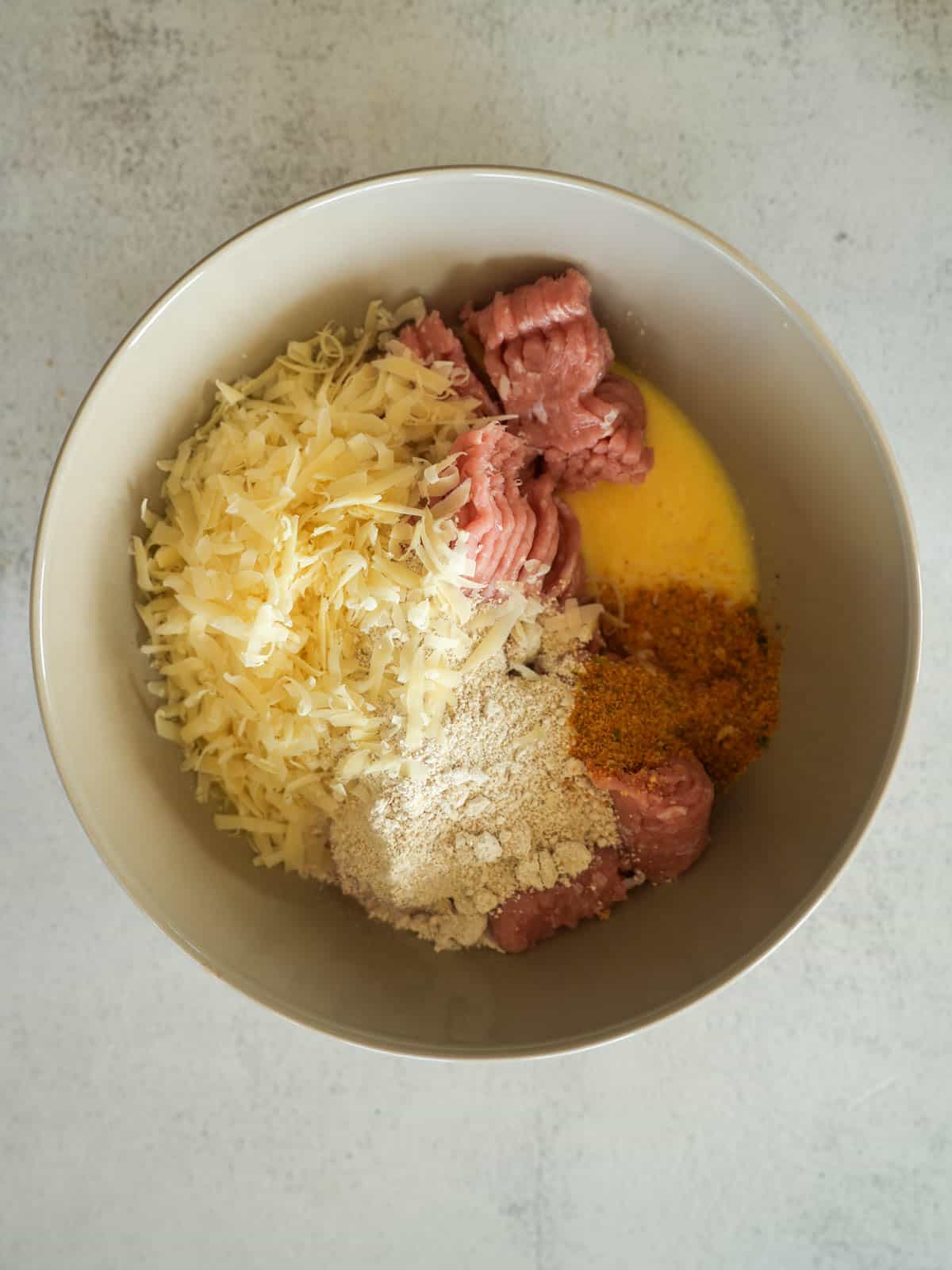 Large mixing bowl containing raw turkey mince, shredded parmesan cheese, oat flour, beaten egg, and garlic and onion powder before mixing.