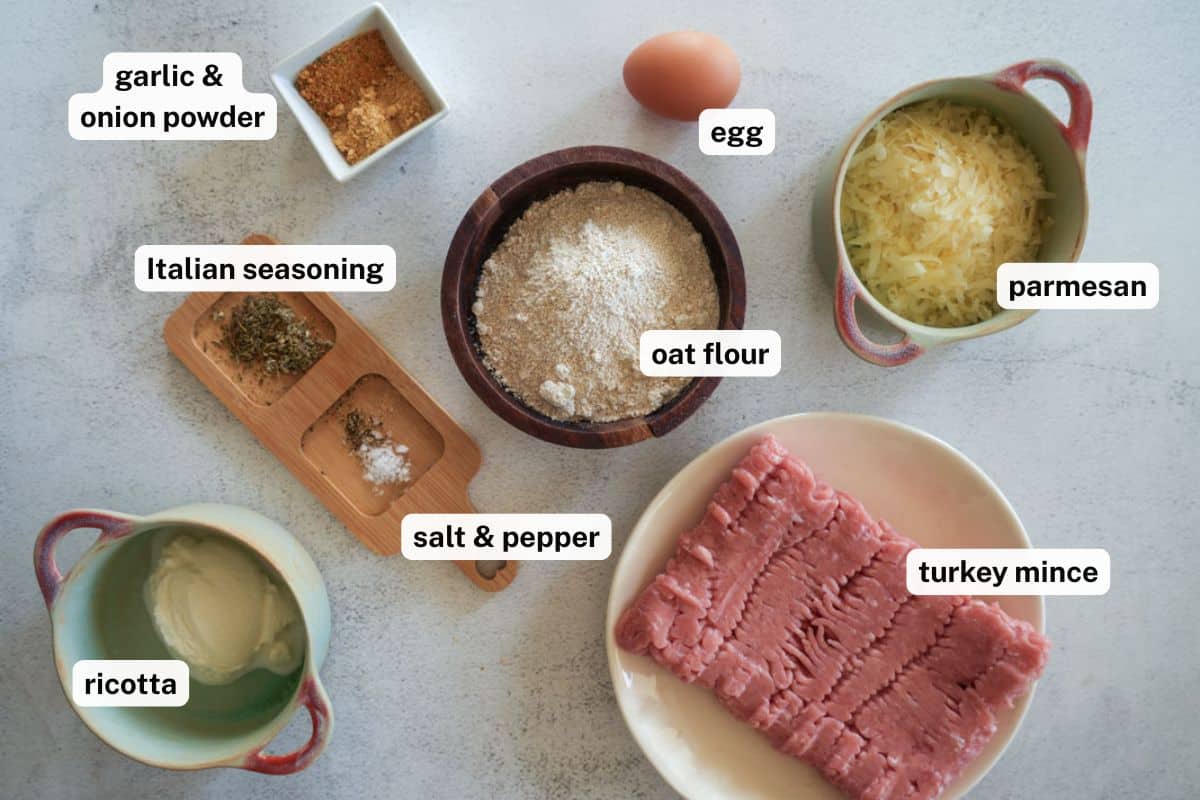 Overhead view of ingredients for air fryer turkey meatballs laid out on a light surface. Items include a small bowl of garlic and onion powder, a brown bowl of oat flour, a whole egg, a small dish of grated parmesan cheese, a plate of raw turkey mince, a small bowl of ricotta cheese, a wooden tray holding Italian seasoning and salt and pepper.