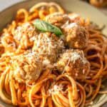 A close-up of a bowl of linguine pasta coated in tomato sauce, topped with golden-brown turkey meatballs, freshly grated Parmesan cheese, and a basil leaf garnish. The food is brightly lit, with a second blurred plate of meatballs in the background.