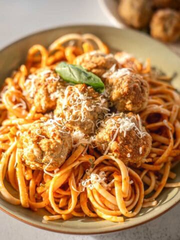A close-up of a bowl of linguine pasta coated in tomato sauce, topped with golden-brown turkey meatballs, freshly grated Parmesan cheese, and a basil leaf garnish. The food is brightly lit, with a second blurred plate of meatballs in the background.