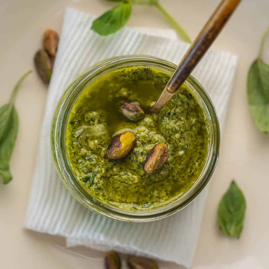 Jar of homemade pistachio pesto topped with whole pistachios, placed on a white plate with basil leaves and lemon in the background.