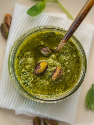 Jar of homemade pistachio pesto topped with whole pistachios, placed on a white plate with basil leaves and lemon in the background.