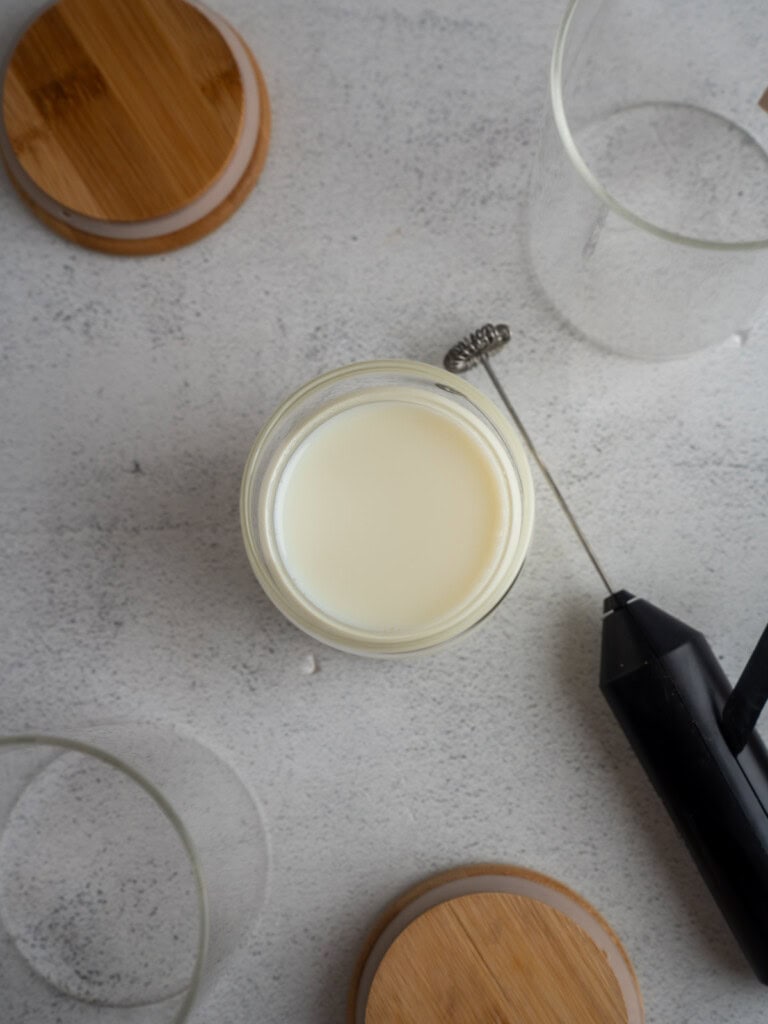Overhead view of a glass of milk on a light gray surface, surrounded by wooden jar lids, empty glass jars, and a small black handheld milk frother.