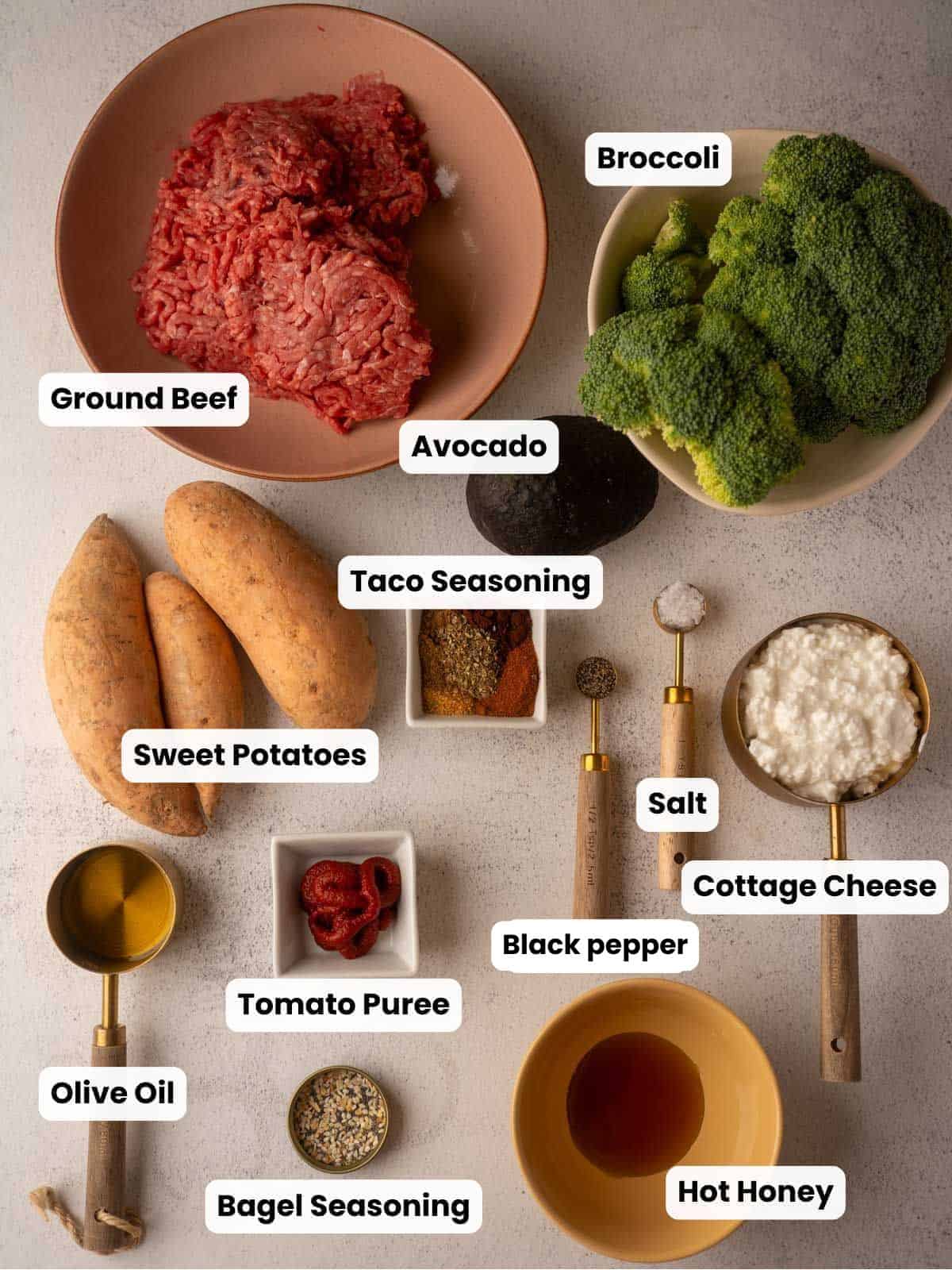 A flat lay photograph of fresh ingredients for a healthy recipe, featuring raw ground beef, sweet potatoes, broccoli, avocado, cottage cheese, assorted spices, and sauces neatly arranged on a light surface.