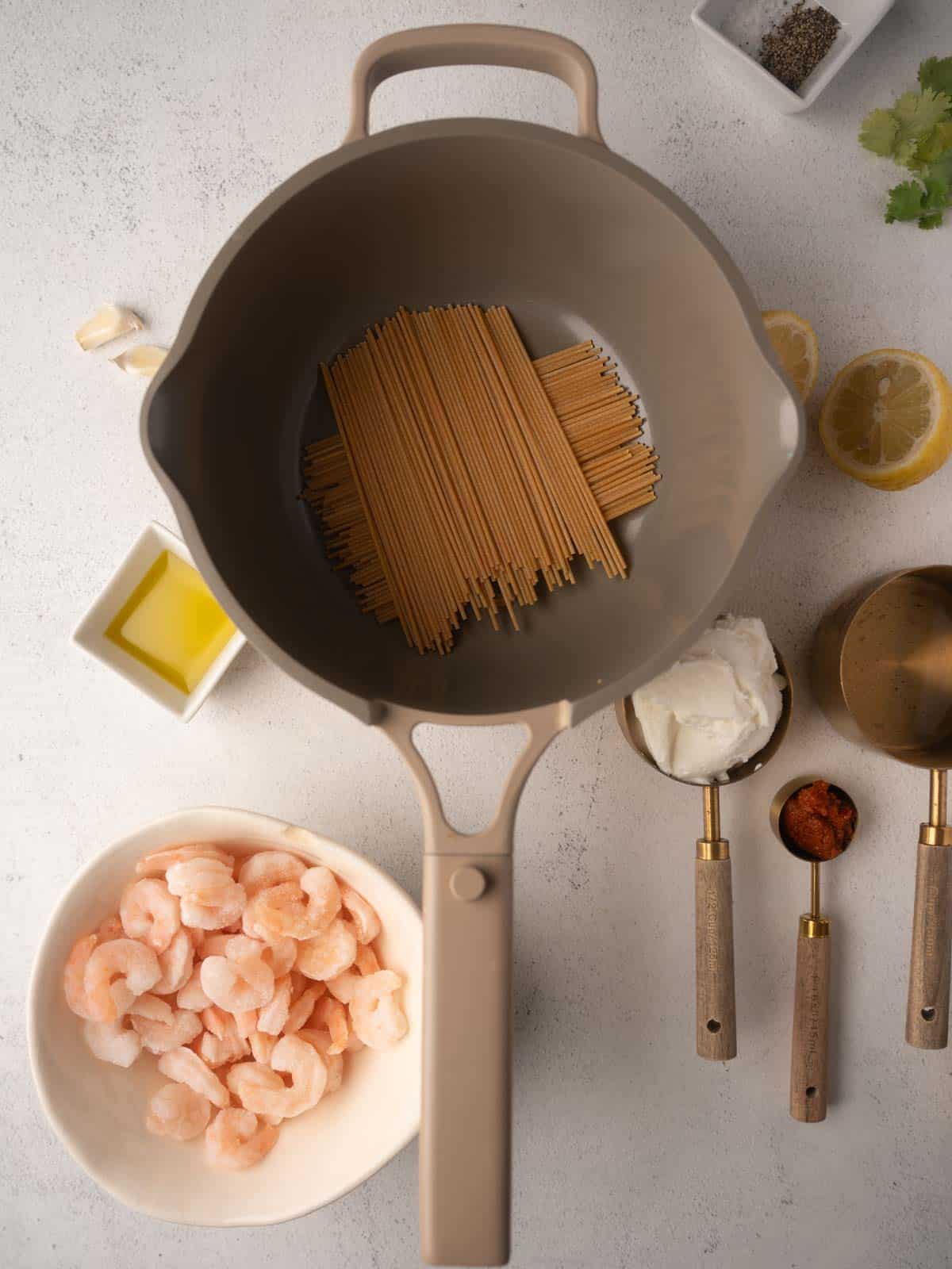 The step 1 for Pesto Ricotta Pasta with Shrimp are arranged on a counter. A pot of dry wholewheat spaghetti is next to a bowl of shrimp, ricotta, and other components, ready to be cooked.