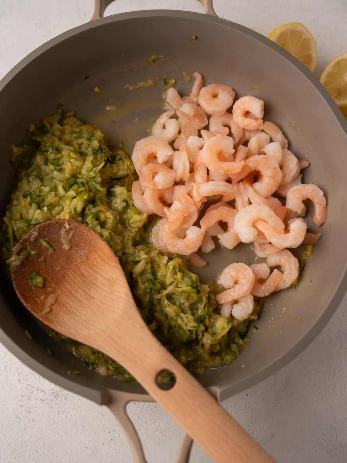 Sautéed grated zucchini and cooked shrimp in a beige nonstick pan, stirred with a wooden spoon. Two lemon halves are visible in the background.