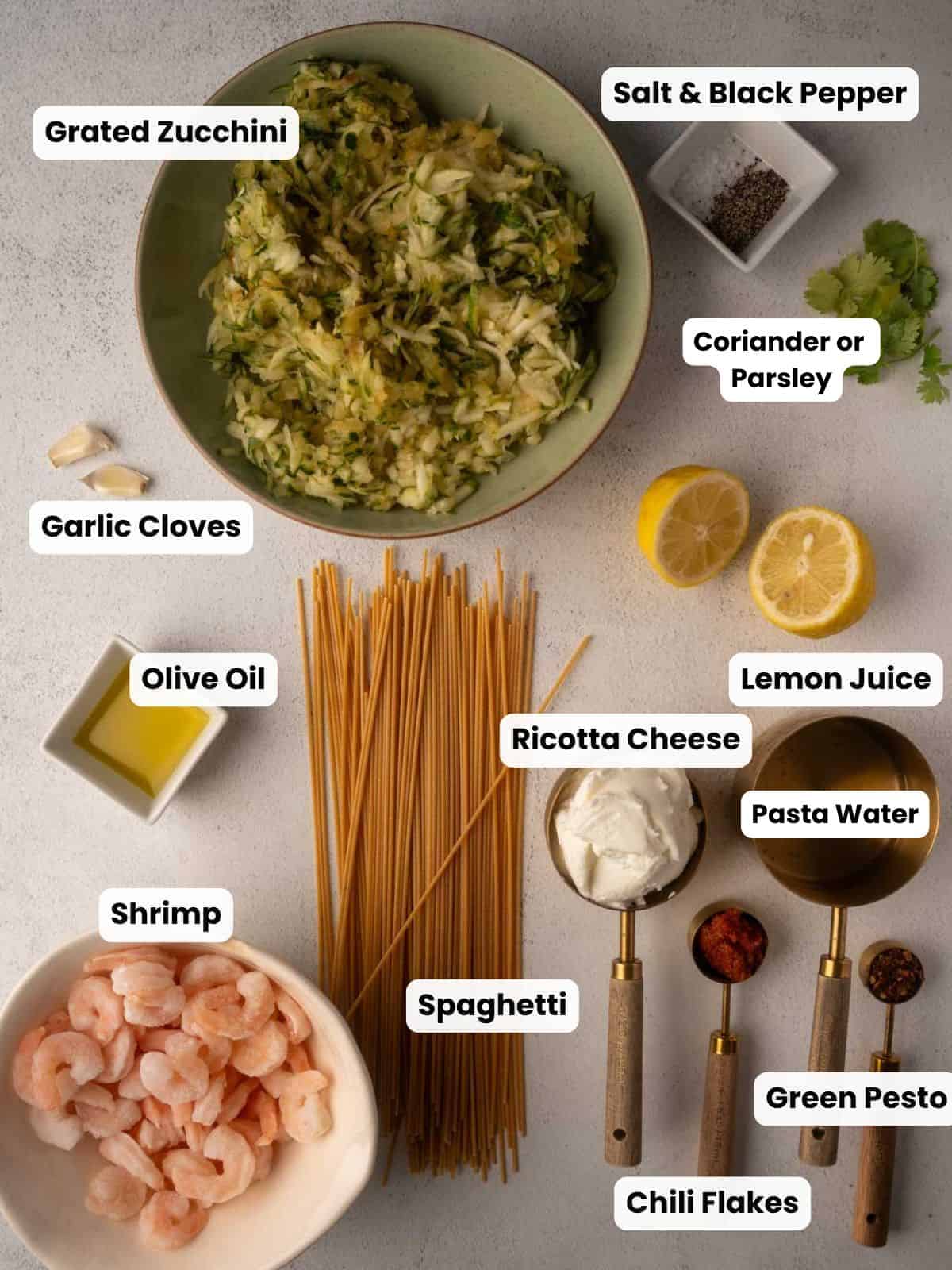 Overhead view of ingredients for pesto ricotta pasta with shrimp. Includes a bowl of grated zucchini, garlic cloves, olive oil, raw shrimp, uncooked spaghetti, small bowls of ricotta cheese, green pesto, pasta water, chili flakes, lemon halves, fresh coriander or parsley, and a dish of salt and black pepper, all arranged neatly on a light surface.