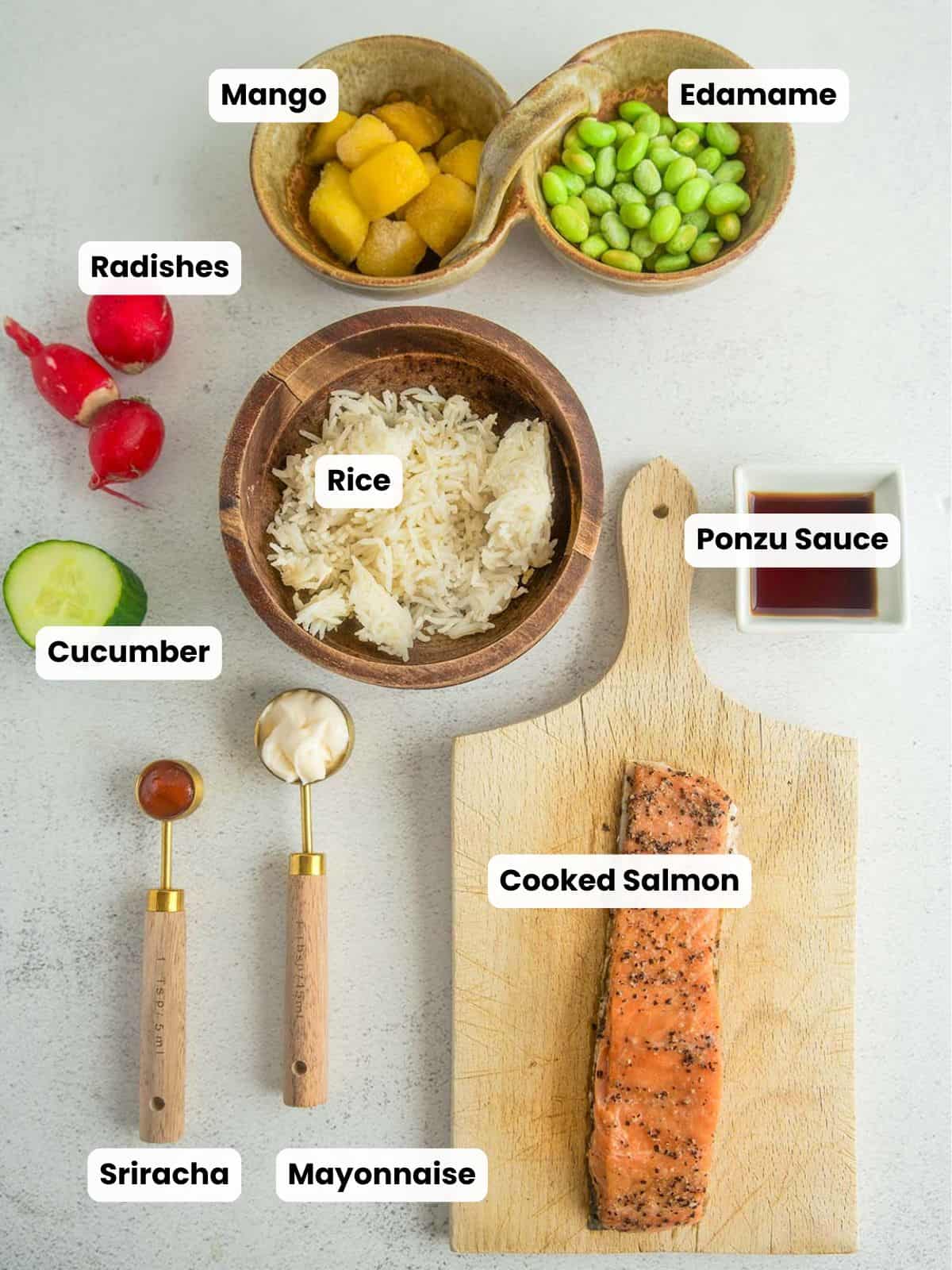 Overhead view of labeled ingredients for a salmon rice bowl, including cooked salmon on a wooden board, rice, mango, edamame, sliced cucumber, radishes, ponzu sauce, mayonnaise, and sriracha arranged on a light surface.