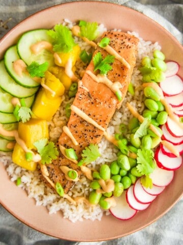 A colorful salmon rice bowl topped with a fillet of seared salmon, sliced cucumbers, mango chunks, radishes, edamame, and cilantro, all drizzled with a creamy sauce and served over white rice in a pink bowl.
