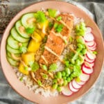 A colorful salmon rice bowl topped with a fillet of seared salmon, sliced cucumbers, mango chunks, radishes, edamame, and cilantro, all drizzled with a creamy sauce and served over white rice in a pink bowl.
