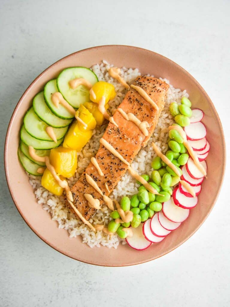 A salmon rice bowl with cucumber, mango, edamame, and radish, drizzled with homemade sriracha mayo sauce.