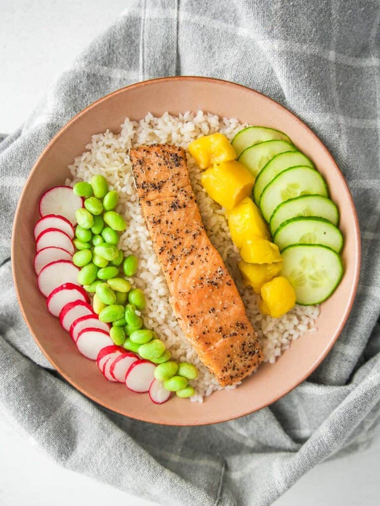 Overhead view of a salmon rice bowl on a gray cloth. A seasoned salmon fillet rests on rice, surrounded by neatly arranged sliced radish, edamame, cubed mango, and sliced cucumber.