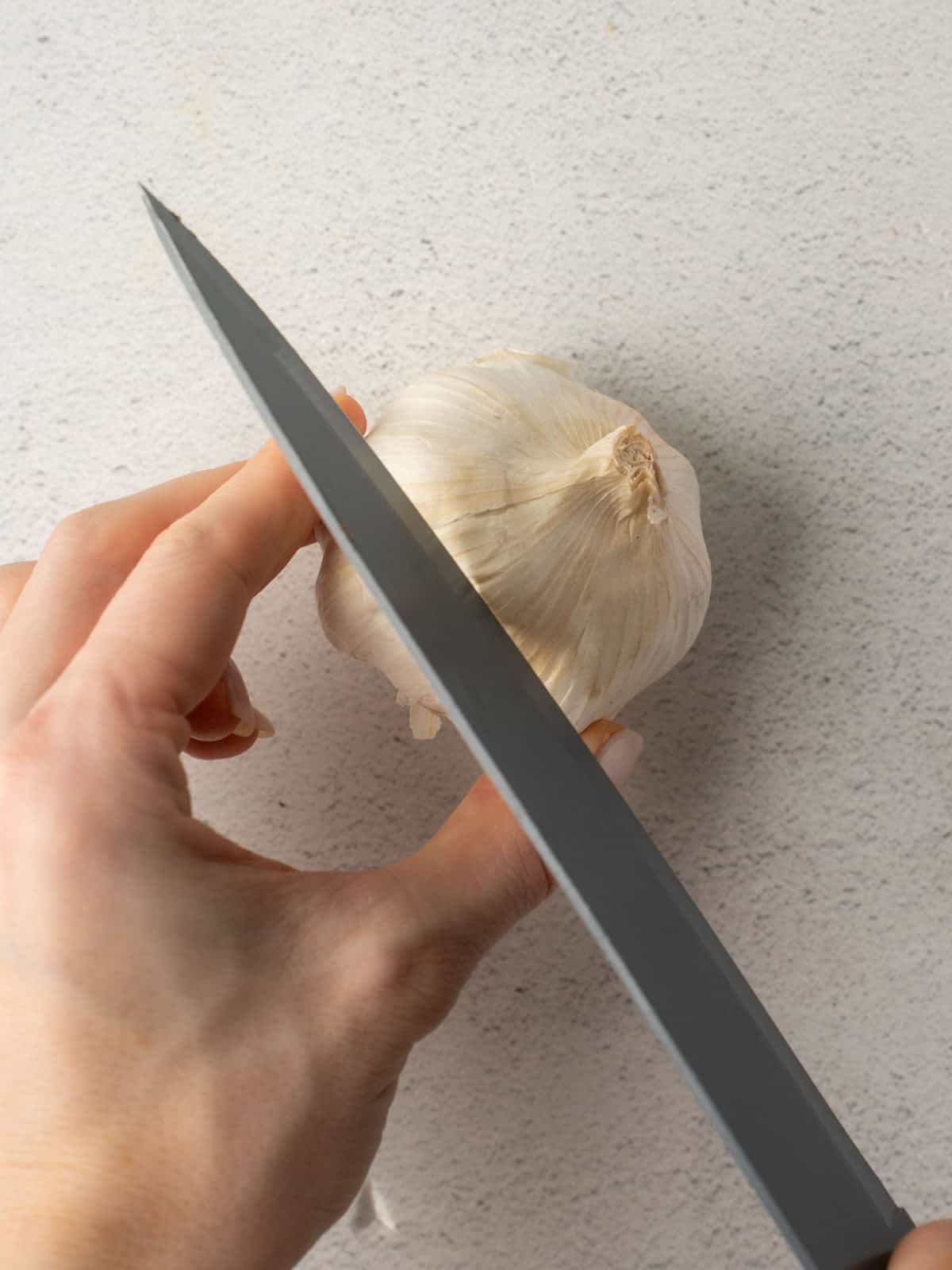 A close-up shot of a hand preparing a garlic bulb by slicing the top, a key step for tomato carrot soup recipe.