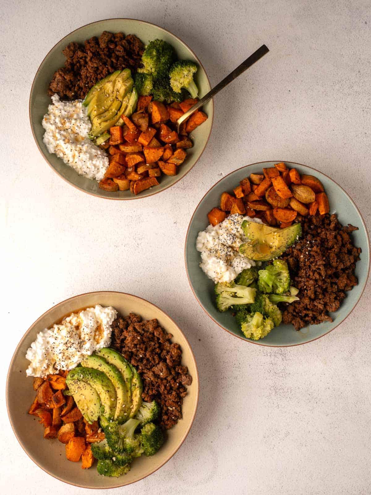 A flat lay photograph of three nutritious protein bowls, each containing lean ground beef, roasted sweet potatoes, broccoli, cottage cheese, and sliced avocado.