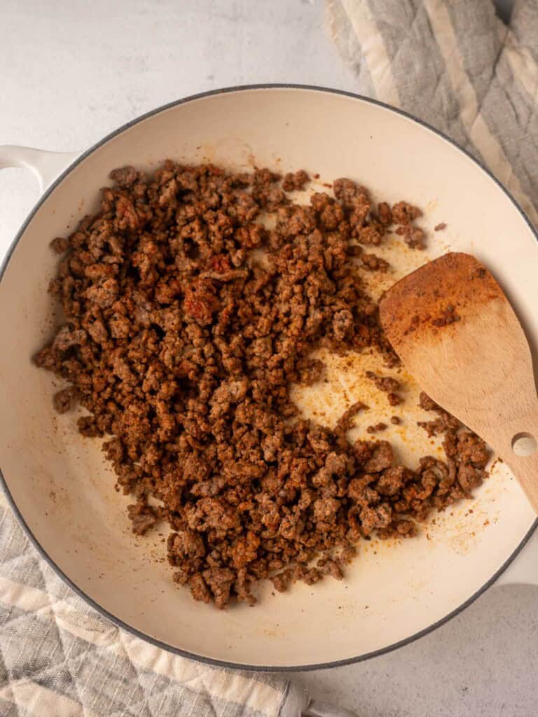 A close-up of lean ground beef, seasoned with taco spices, being cooked and crumbled with a wooden spatula in a light-colored skillet.